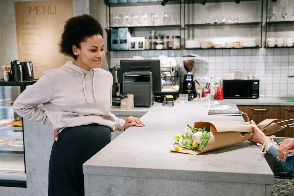 A young woman with natural curly hair, wearing a light grey hoodie and dark trousers, stands on one side of a white marble counter in a modern café or flower shop. She is observing a floral gift bein