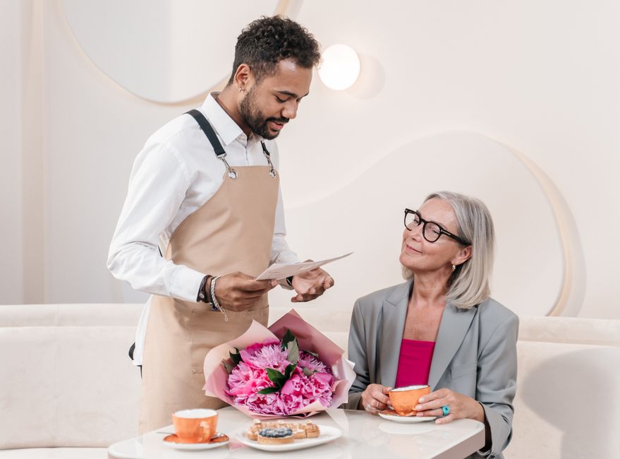 A flower delivery florist from bowesparkflowers.co.uk presents a vibrant bouquet of pink peonies and roses wrapped in pink paper with green foliage to a smiling elderly woman with grey hair, glasses, 