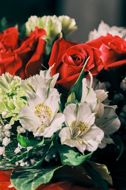 A floral bouquet featuring red roses, white lilies with pale yellow and dark purple markings, and green foliage arranged in a lush, rounded style. The flowers are fresh, with velvety petals and sturdy