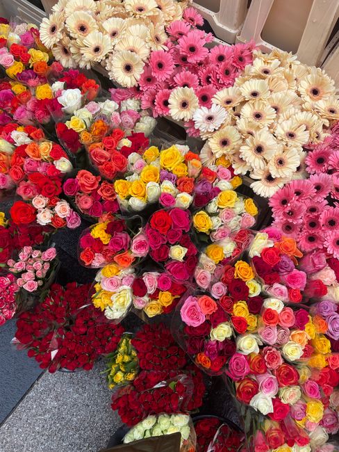 A vibrant display of fresh floral arrangements featuring a variety of roses, gerbera daisies, and other blooms in shades of pink, red, yellow, and cream. The bouquets are densely packed, showcasing di